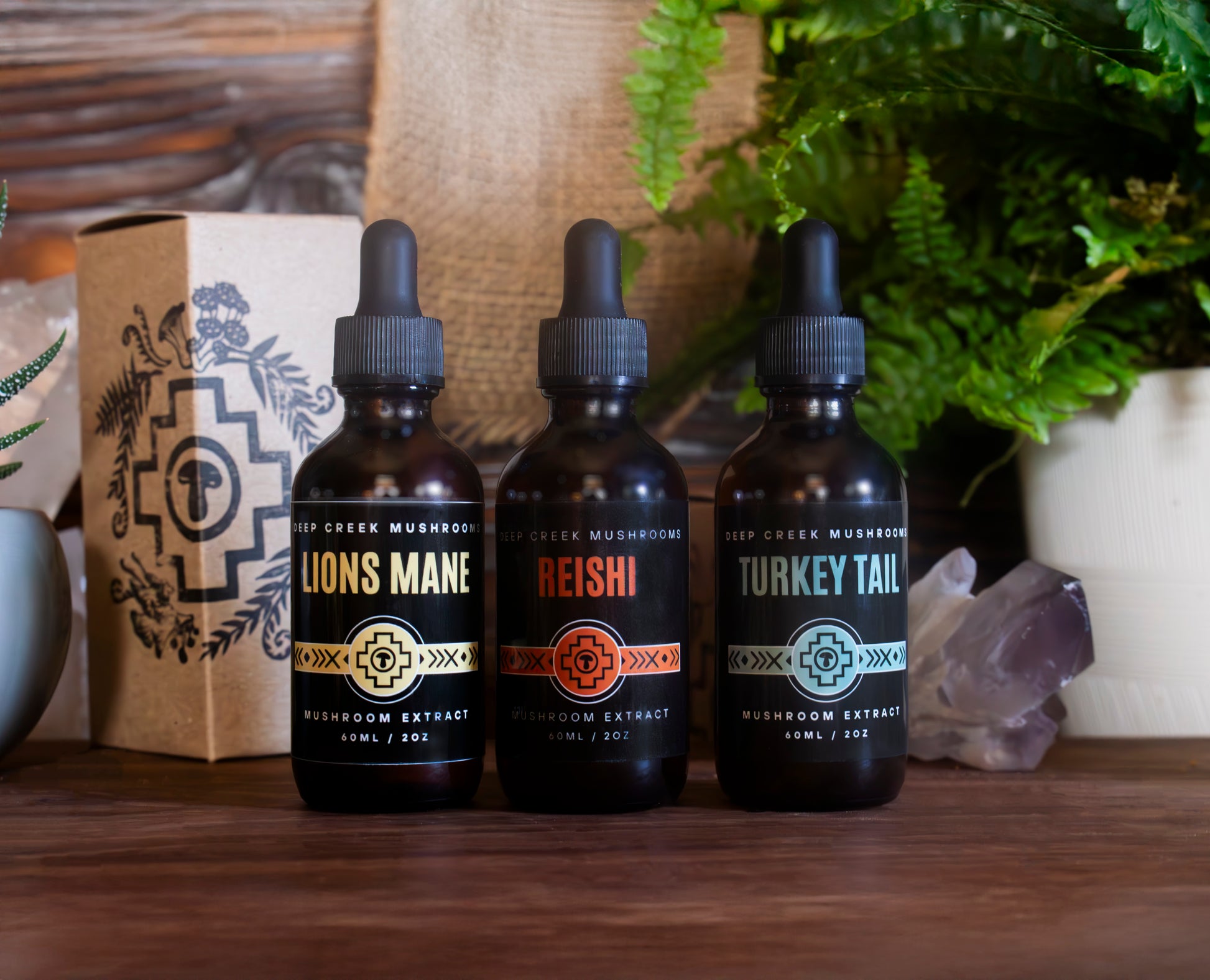 Three bottles of mushroom extracts from Deep Creek Mushrooms labeled 'Lions Mane', 'Reishi', and 'Turkey Tail' on a wooden surface with a plant and crystal in the background.